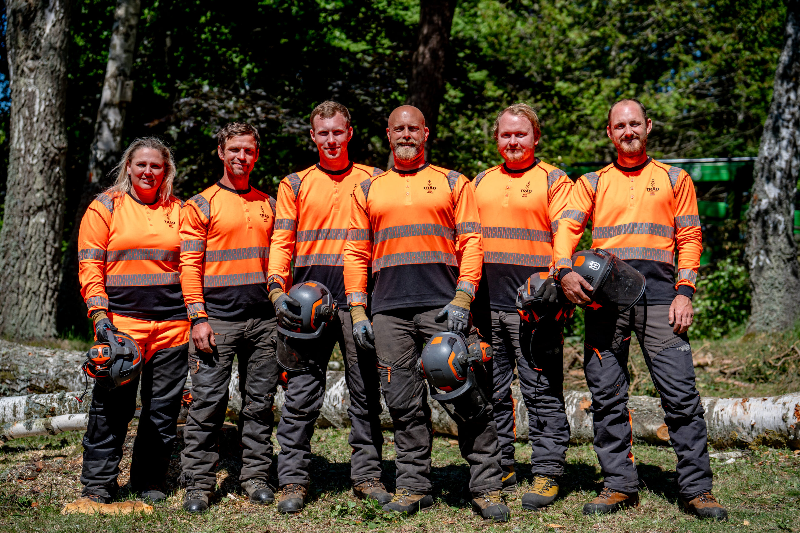 Sex personer i orange varselkläder står på rad i skogen med hjälmar i händerna